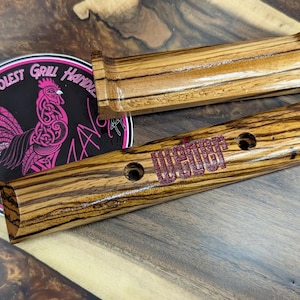 May include: Two wooden grill handles with a zebrawood grain pattern. One handle has the word "WEBER" in red lettering. A circular sticker with a pink rooster and the text "THE COOLEST GRILL HANDLE MAN" is also visible.
