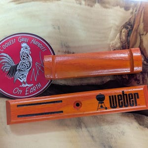 May include: An overhead shot of two orange-stained wooden grill handles and a red circular emblem. The emblem reads "The Coolest Grill Handles On Earth" and features a rooster illustration. One handle has the "Weber" logo.
