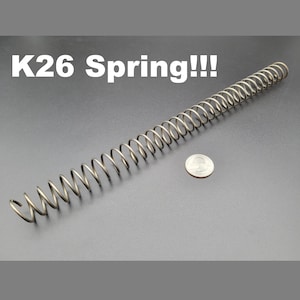 May include: A silver metal spring, approximately 6 inches long, is laid out on a gray surface. A silver US quarter is placed next to the spring for size comparison. The text "K26 Spring!!" is written in white letters on a black background above the spring.