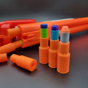 May include: Three orange and white Nerf darts with different coloured tips. The tips are green, blue, and grey. The darts are sitting on a grey surface.