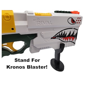 May include: A white and green Nerf Rival Kronos blaster with a black stand. The blaster has a yellow and black design and a shark graphic on the side. The text "NERF RIVAL" is printed on the blaster.