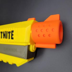 以下が含まれることがあります： 側面には「TNITE」の文字がプリントされた、黄色とオレンジ色の玩具銃。銃には「SMHHH...」の文字が入ったグレーの部分と、円形のディテールが施されたオレンジ色の銃身があります。おもちゃは灰色の背景に配置されています。