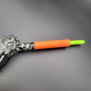 May include: A toy gun with a camouflage pattern on the body. The barrel has an orange sleeve with small holes and a green tip. The toy is set against a dark gray background.
