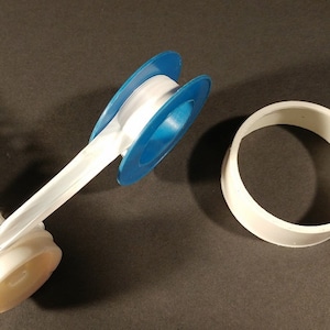 May include: A collection of plumbing components, including a white and blue Teflon tape dispenser, a white plastic ring, and a spring-loaded mechanism. The items are arranged on a dark surface.