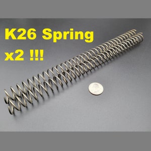 May include: Two silver metal springs, each about 6 inches long, are laid out side-by-side on a gray surface. A US quarter is placed below the springs for scale. The text "K26 Spring x2!!" is written in yellow letters at the top of the image.