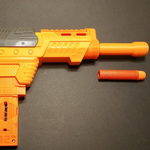 Op de afbeelding: Een oranje en grijze speelgoedblaster met een afneembare oranje dart. De blaster heeft een magazijn en een loop. De dart is cilindrisch met een oranje punt. Op een zwart oppervlak.