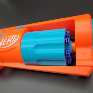 May include: A close-up of an orange and blue toy gun with the word "NERF" printed on the side. The gun has a blue cylinder with a purple and blue rotating drum.