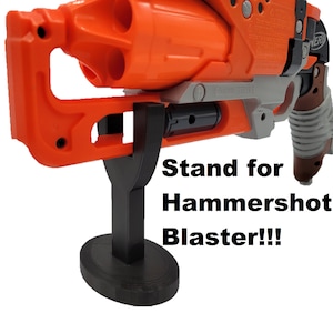 May include: A black plastic stand for a Nerf Hammershot blaster. The stand has a rectangular base and a vertical support. The stand is designed to hold the blaster upright.