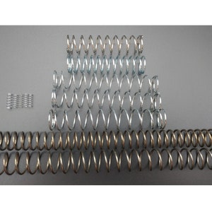 May include: An assortment of metal coil springs in various sizes and colors, including silver and bronze. The springs are arranged on a gray surface, showcasing their different lengths and thicknesses. A small group of tightly wound springs is also present.