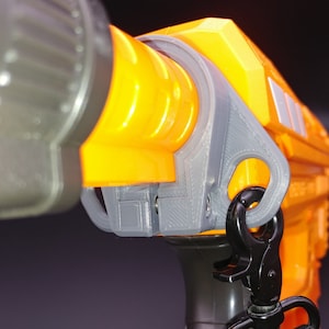 May include: Close-up of a bright orange and gray toy gun with a black strap attached. The gun has a textured surface and a gray trigger guard.