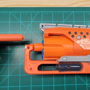May include: An orange and gray toy gun with a black barrel and a blue and orange cylinder. The gun has a silver trigger and a black handle. The gun is on a green cutting mat with a ruler.