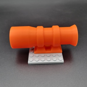 Puede incluir: Un cañón de juguete de plástico naranja, con un cañón cilíndrico y dos bandas elevadas. El cañón está montado sobre una pequeña base rectangular hecha de bloques grises entrelazados. El juguete está diseñado para jugar y exhibir.