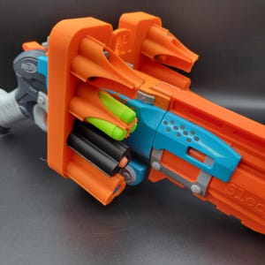 Puede incluir: Un juguete de blaster Nerf en naranja, azul y gris. La pistola de juguete tiene múltiples cañones naranjas y una sección azul con diseños circulares. El blaster incluye munición verde y negra. El mango está envuelto en cinta blanca.