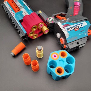 Puede incluir: Un lanzador de juguete Zuru X-Shot con un cañón turquesa y naranja y un cilindro giratorio rojo, acompañado de un cilindro azul separado, tres casquillos naranjas y un casquillo amarillo con punta gris sobre una superficie oscura.