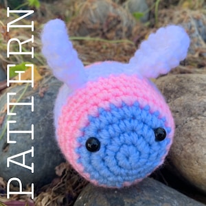 Puede incluir: Un juguete de amigurumi tejido a crochet en forma de abeja con rayas rosas y azules. La abeja tiene dos antenas blancas y ojos negros.