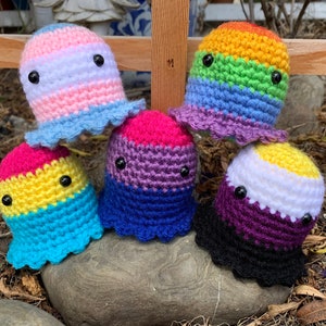 Peut inclure: Cinq créatures crochetées à la main, chacune avec un schéma de couleurs différent représentant les drapeaux de la fierté LGBTQ+. Les créatures ont des yeux noirs en forme de bouton et des bords froncés. Elles sont présentées sur un rocher en extérieur.