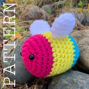 Puede incluir: Un juguete de abeja de crochet con rayas rosas, amarillas y azules. La abeja tiene alas blancas y ojos negros.