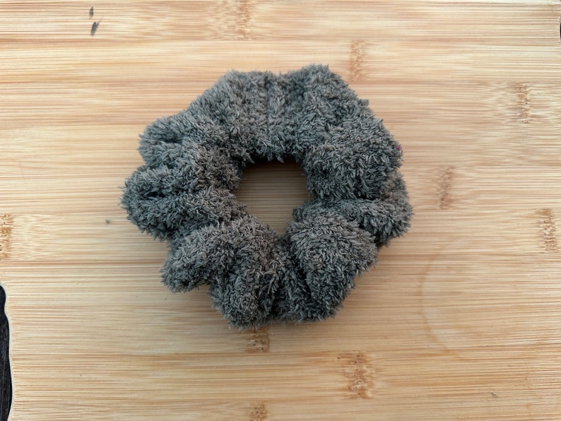 May include: A gray fuzzy scrunchie hair tie. The scrunchie is made of a soft, plush fabric.