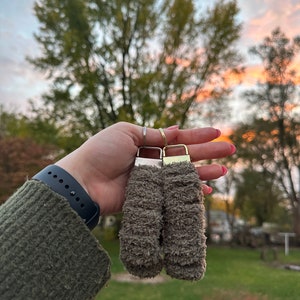 May include: Two fuzzy brown keychains with gold rings. The keychains are hanging from the rings and are in the shape of a long, thin rectangle.