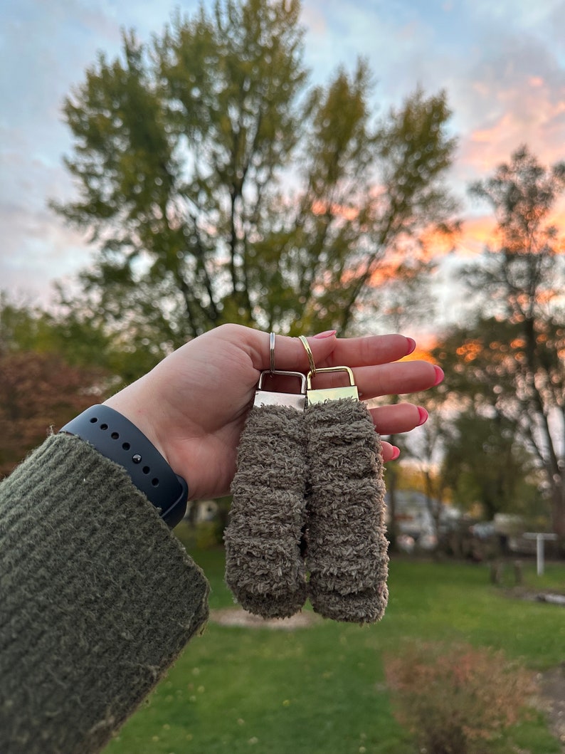 May include: Two fuzzy brown keychains with gold rings. The keychains are hanging from the rings and are in the shape of a long, thin rectangle.