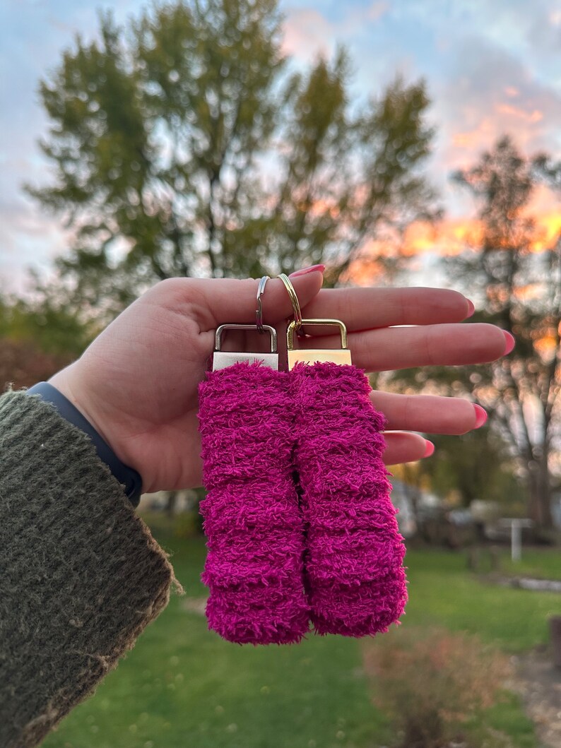 May include: Two pink fuzzy keychains with silver and gold metal rings. The keychains are made of a soft, fluffy material.