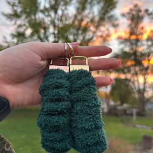 May include: Two green fuzzy keychains with gold metal key rings. The keychains are made of a soft, plush material.