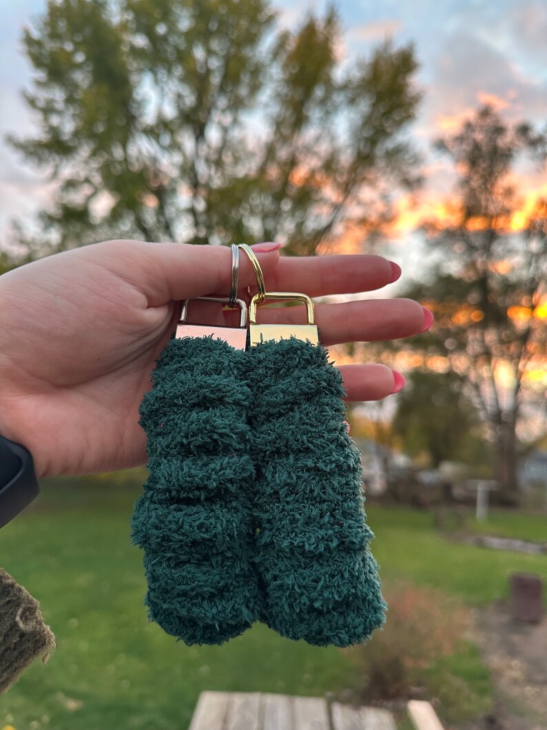 May include: Two green fuzzy keychains with gold metal key rings. The keychains are made of a soft, plush material.
