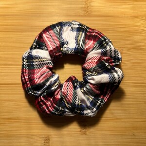 May include: A plaid scrunchie in red, black, white, and blue. The hair accessory is circular and made of fabric. The scrunchie is on a wooden surface.