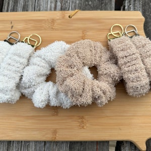 May include: Three fluffy keychains in white and beige. The keychains are made of a soft, fuzzy material and have a metal ring attached to them.