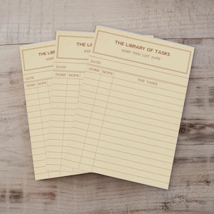 May include: Three cream-coloured paper task pads with brown ink. Each pad features the header THE LIBRARY OF TASKS, KEEP THIS LIST SAFE, and columns labelled DATE, DONE, NOPE, and THE TASKS with horizontal lines for writing entries.