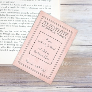 May include: A pink card with the title "The Witch's Curse: An Operatic Tragedy." The card states it's a play written by Jo March and set by The March Sisters, dated December 25th, 1862. The card is placed on a wooden surface, alongside a book.