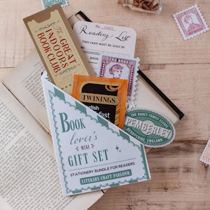 A stationery gift set for readers styled on a wooden tabletop with an open vintage book. The set is packaged in a blind date with a book style pocket and reads 'Book lover's mini GIFT SET' on the front. It has a tan ticket-style bookmark, a reading list card, a vintage-style stamp sticker, a Pemberley travel sticker and a Twinings tea packet tucked inside.