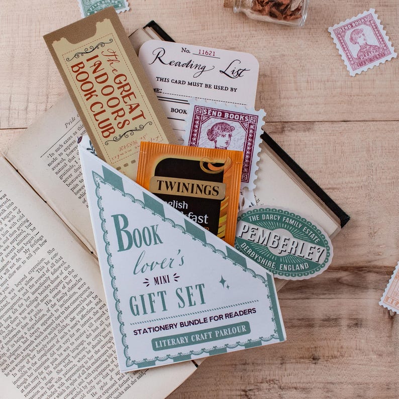 A stationery gift set for readers styled on a wooden tabletop with an open vintage book. The set is packaged in a blind date with a book style pocket and reads 'Book lover's mini GIFT SET' on the front. It has a tan ticket-style bookmark, a reading list card, a vintage-style stamp sticker, a Pemberley travel sticker and a Twinings tea packet tucked inside.