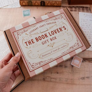 May include: A rectangular cardboard gift box with a cream and light pink striped design. The top features decorative brown text reading LITERARY CRAFT PARLOUR THE BOOK LOVER'S GIFT BOX Stationery & treats for readers, with ornate flourishes.