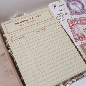 May include: A cream-colored task notepad with brown text reading THE LIBRARY OF TASKS and KEEP THIS LIST SAFE, featuring columns for DATE, DONE, NOPE, and THE TASKS. Beside it are vintage-style library cards and stamps with red and purple ink.