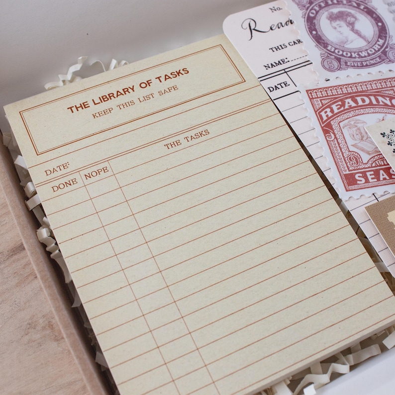 May include: A cream-colored task notepad with brown text reading THE LIBRARY OF TASKS and KEEP THIS LIST SAFE, featuring columns for DATE, DONE, NOPE, and THE TASKS. Beside it are vintage-style library cards and stamps with red and purple ink.