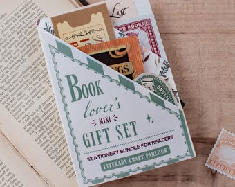 May include: A triangular paper envelope labeled Book lover's MINI GIFT SET STATIONERY BUNDLE FOR READERS LITERARY CRAFT PARLOUR containing various paper items, including a tan ticket, a red stamp, and a floral card, resting on an open book.