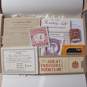 May include: A collection of paper stationery items in a box, including a Library of Tasks card, a Reading List card, a Netherfield Ball invitation, a Great Indoors Book Club ticket, a book-shaped pin, and a Twinings tea bag with vintage graphics.