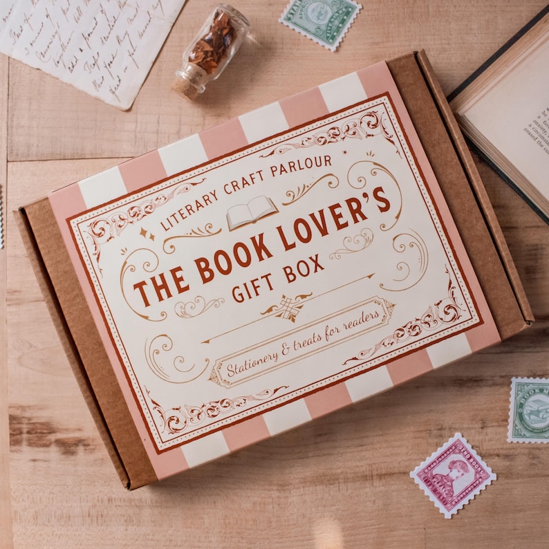 A flat-lay view of a 'The Book Lover's Gift Box' by Literary Craft Parlour, placed on a wooden table with vintage props like old books, stamps, and a letter. The packaging is similar to a vintage chocolate or cookie box or tin and has a pink and white striped background with red text and gold accents.