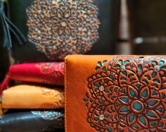 May include: A stack of leather wallets in various colors, including black, red, and tan. The wallets feature a detailed, carved mandala design. The front-most wallet is tan with a turquoise interior and silver studs.