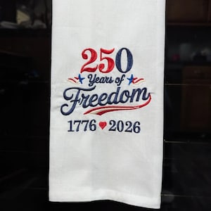 May include: White kitchen towel with embroidered text and design. The text reads "250 Years of Freedom" with the years 1776 and 2026. The design includes red, white, and blue stars and a heart.