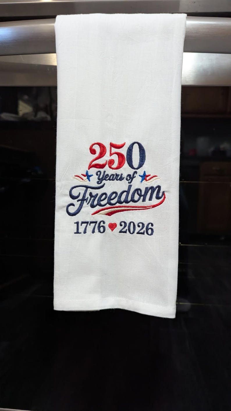 May include: White kitchen towel with embroidered text and design. The text reads "250 Years of Freedom" with the years 1776 and 2026. The design includes red, white, and blue stars and a heart.