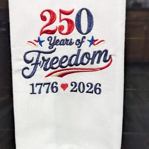 May include: White towel embroidered with the text "250 Years of Freedom" in red and blue. The numbers "1776" and "2026" are also embroidered in blue, with a red heart symbol between them. The towel is a patriotic item.