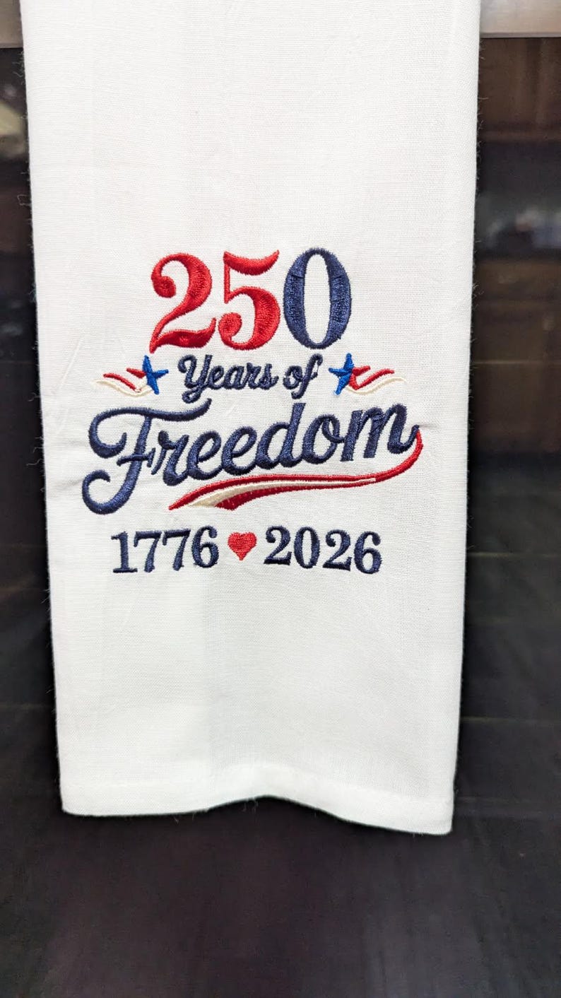 May include: White towel embroidered with the text "250 Years of Freedom" in red and blue. The numbers "1776" and "2026" are also embroidered in blue, with a red heart symbol between them. The towel is a patriotic item.