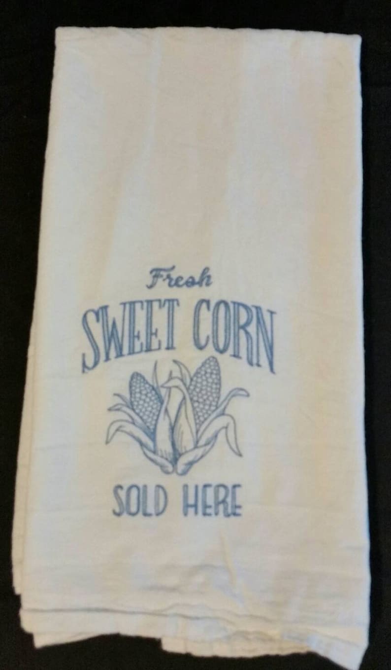 May include: White kitchen towel with blue embroidery that reads "Fresh Sweet Corn Sold Here". The embroidery also includes a graphic of two ears of corn.