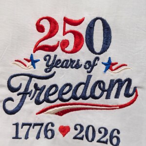 May include: White fabric embroidered with the text "250 Years of Freedom" in red and blue thread. The numbers "250" are in red and blue, with the years "1776" and "2026" below. A red heart is embroidered between the years.
