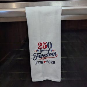 May include: White kitchen towel with the text "250 Years of Freedom 1776-2026" embroidered in red and blue. The towel is hanging on an oven door. The oven is stainless steel.