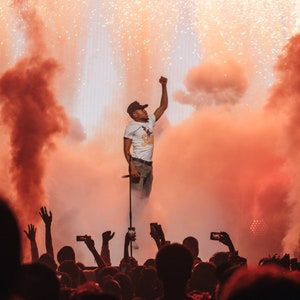May include: A live concert scene with a performer on stage, raising their fist. The stage is filled with orange smoke and pyrotechnics. The audience below is holding up phones, capturing the moment.
