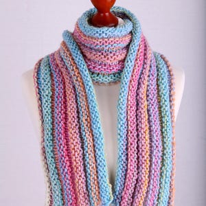 Könnte beinhalten: Ein bunter gestreifter Strick Schal in Blautönen, Rosa und Orange. Der Schal wird auf einer weißen Schaufensterpuppe präsentiert.