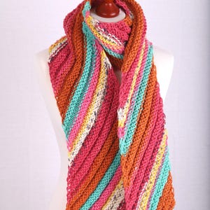 Könnte beinhalten: Ein farbenfroher, handgestrickter Schal mit diagonalen Streifen in Rosa, Orange, Türkis, Gelb und Weiß. Der Schal wird auf einer Schaufensterpuppe präsentiert und zeigt sein strukturiertes, warmes Design. Ein gemütliches Accessoire für kaltes Wetter.